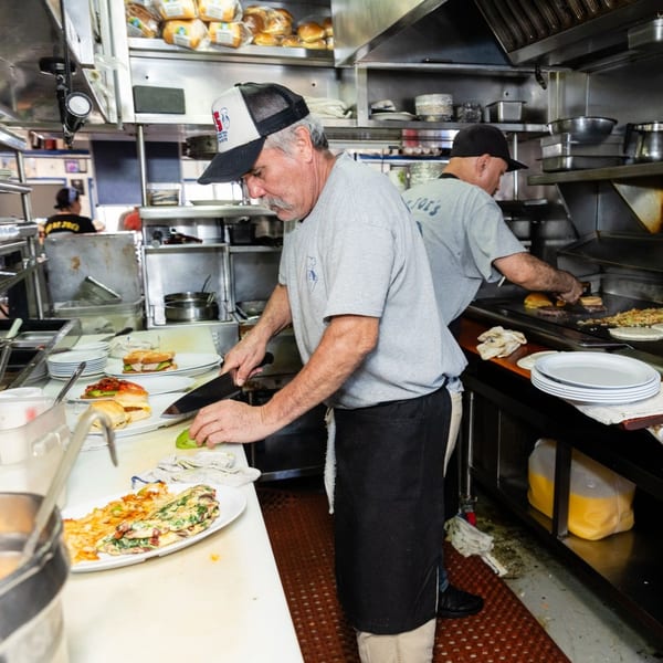 Two chefs in a busy diner kitchen, one slicing avocado, the other cooking on a griddle.