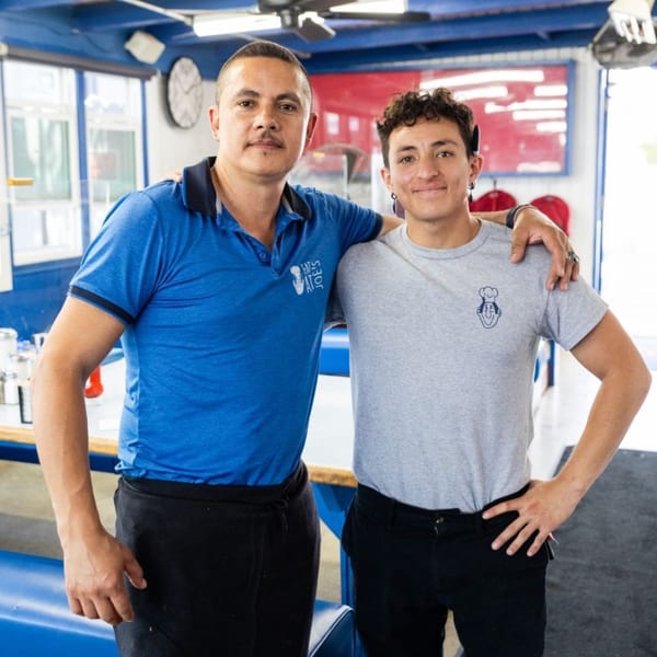 Two men, one in a blue Eat at Joe's shirt, stand smiling in a diner.