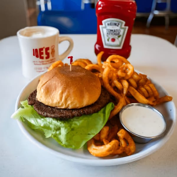 A burger with lettuce, curly fries, dipping sauce, and a "Joe's" mug on a diner table.