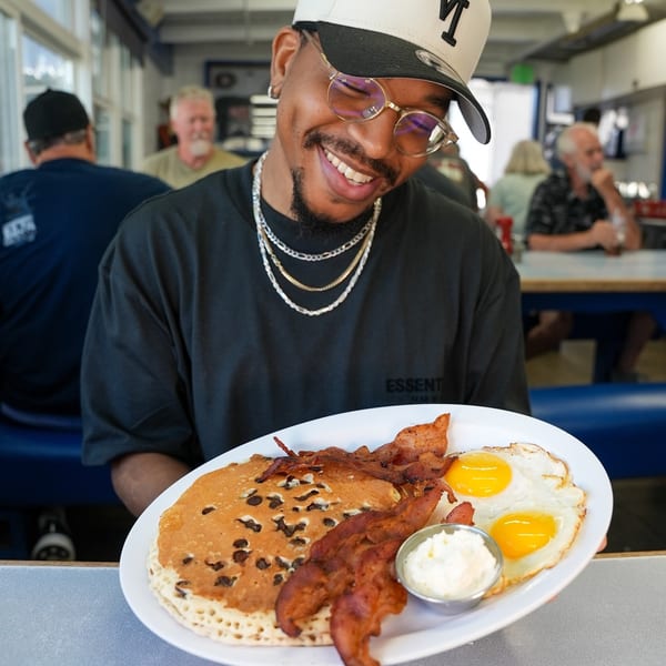 Smiling man in a diner holding a plate of chocolate chip pancakes, bacon, and fried eggs.