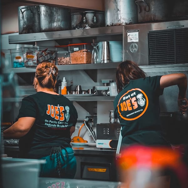 Two Eat at Joe's diner staff members working in a busy kitchen with large pots and containers.