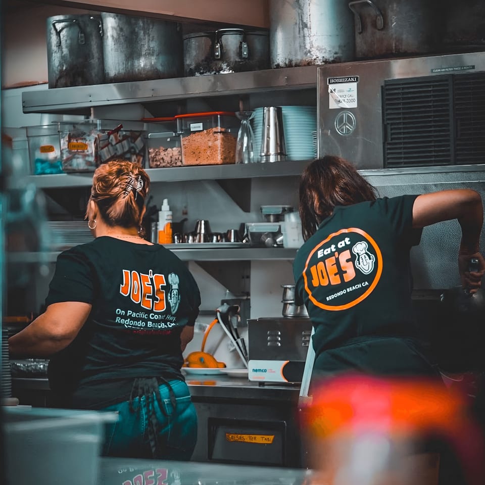 Two Eat at Joe's diner staff members working in a busy kitchen with large pots and containers.