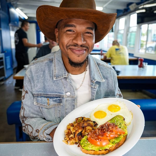 Man in a cowboy hat and denim jacket holding a diner breakfast plate with eggs and avocado toast.