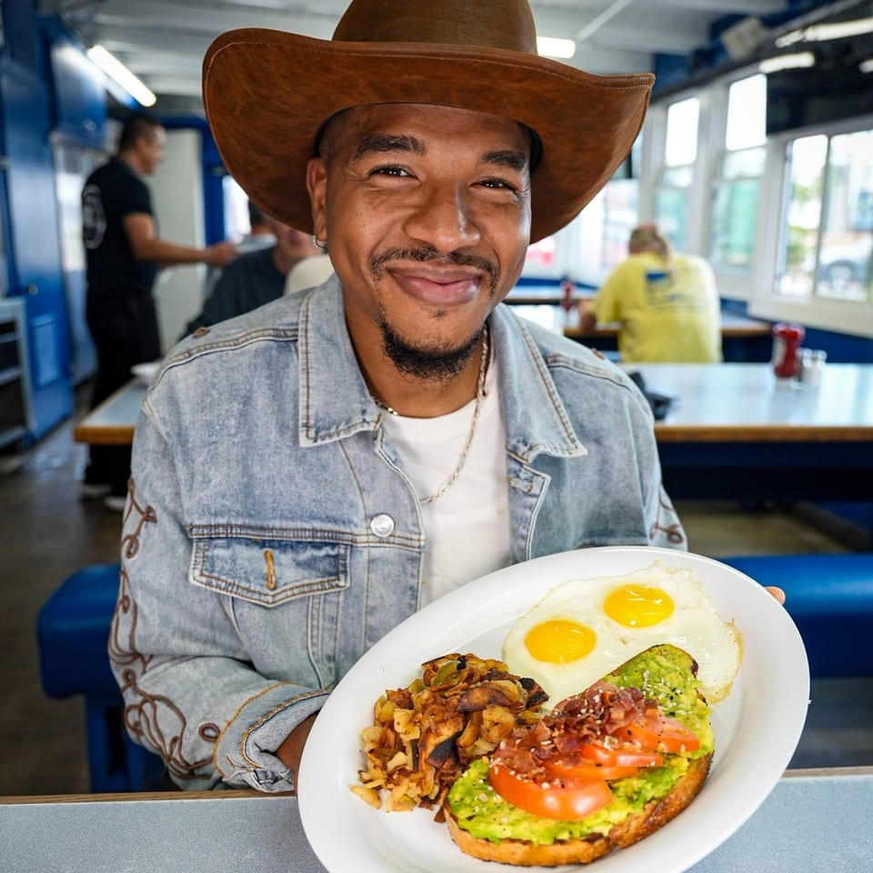 Man in a cowboy hat and denim jacket holding a diner breakfast plate with eggs and avocado toast.