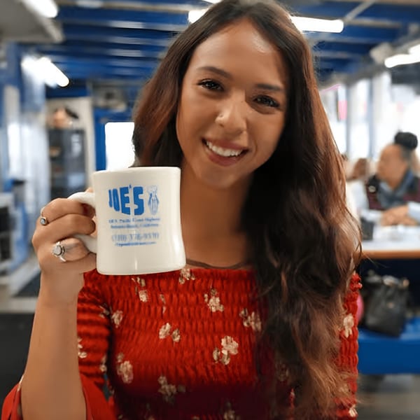 Smiling woman in a red dress holding a white "Eat at Joe's" diner mug.