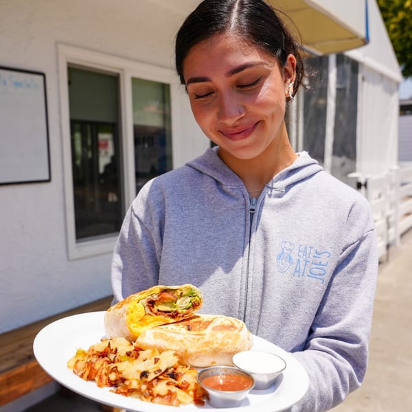 A smiling server in an "Eat at Joe's" hoodie holds a breakfast burrito with hash browns and sauces.